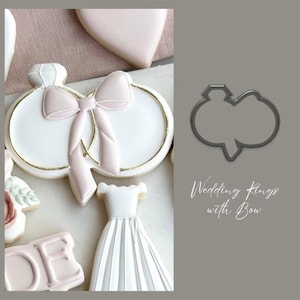 May include: A cookie cutter in the shape of two wedding rings with a bow on top. The text "Wedding Rings with Bow" is written below the cutter.