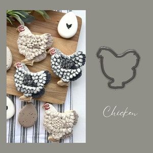 May include: Four decorated sugar cookies shaped like chickens, with a cookie cutter in the shape of a chicken. The cookies are decorated with white, black, and gray icing, and the cookie cutter is gray. The text "Chicken" is written in cursive on the right side of the image.