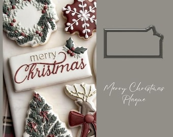 May include: Christmas-themed cookies, including a wreath, snowflake, Christmas tree, and reindeer, are arranged on a white surface. A rectangular cookie with "Merry Christmas" written in red script is also present. A gray plaque is on the right.