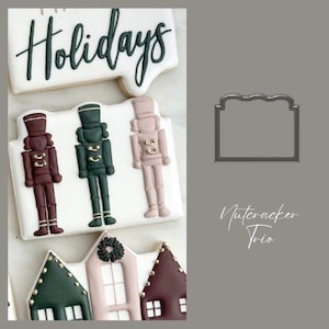 May include: Three gingerbread nutcracker cookies decorated with green, red and white icing. The cookies are arranged in a row on a white background. The text "Holidays" is visible in the background. A metal cookie cutter in the shape of a rectangle is also visible.