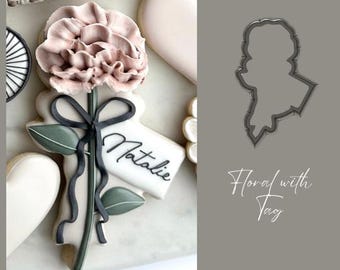 May include: A cookie decorated with a pink flower, green leaves, a black ribbon, and a white tag that says "Natalie". The cookie is on a white surface. A cookie cutter in the shape of a flower with a tag is shown in the background.
