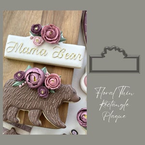 May include: A cookie cutter in the shape of a rectangle with a floral design on the top edge. The text "Floral Thin Rectangle Plaque" is below the cutter.