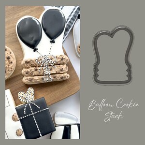 May include: A cookie cutter in the shape of a stack of cookies with two balloons on top. The cookie cutter is made of metal and is gray in color. The text "Balloon Cookie Stack" is written below the cookie cutter.