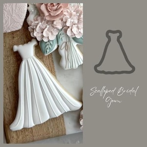 May include: A white fondant cookie decorated to look like a bridal gown. The cookie is on a wooden surface. The text "Scalloped Bridal Gown" is written in cursive on a gray background.