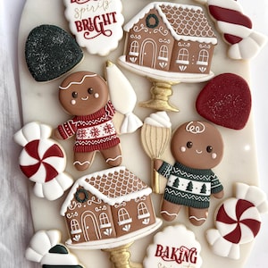 May include: A selection of festive Christmas cookies on a white surface. The cookies feature gingerbread men, gingerbread houses on golden stands, candy canes, and cookies with the text "Baking Spirits Bright" in red script.