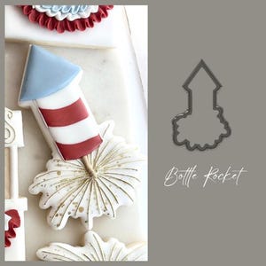 May include: A decorated cookie in the shape of a red, white, and blue rocket with a gold-colored burst. The cookie is part of a set of patriotic-themed treats. The words "Bottle Rocket" are written in cursive.