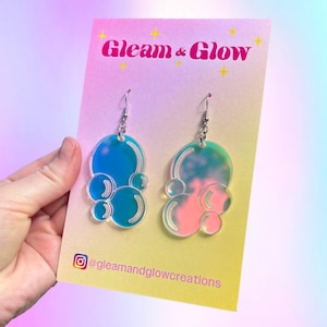 May include: Iridescent bubble-shaped earrings with silver hooks. One earring is blue, the other pink, both with a gradient effect. The earrings are displayed on a card with the text "Gleam & Glow" and an Instagram handle.