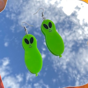 May include: A pair of green alien-shaped earrings with black eyes and a smiling face. The earrings are hanging from silver hooks.