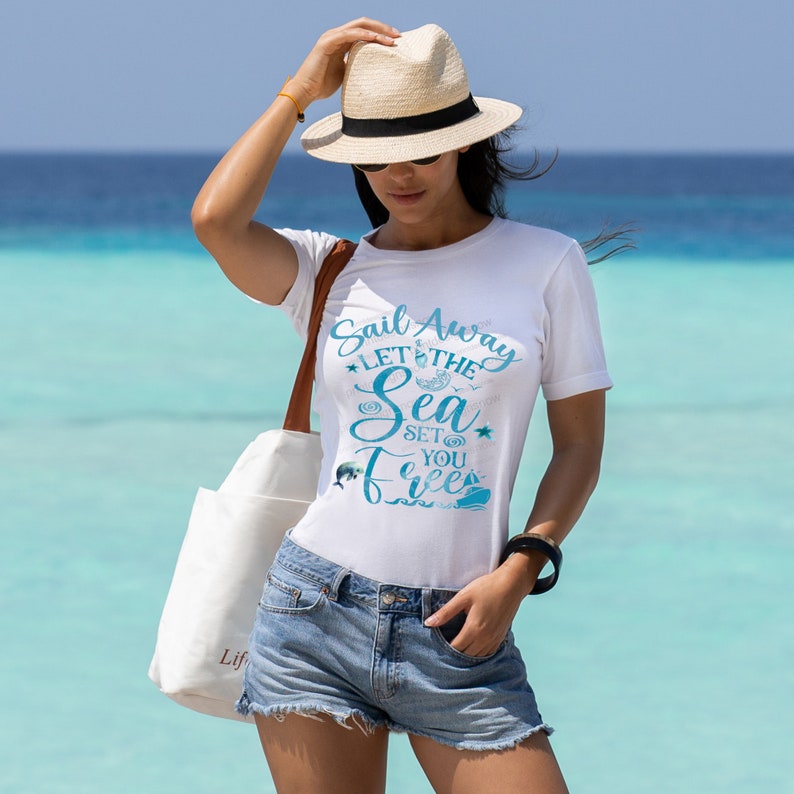 May include: A white t-shirt with a blue graphic that says "Sail Away Let the Sea Set You Free". The graphic includes a sailboat, a dolphin, and a starfish. The shirt is being worn by a person standing on a beach.