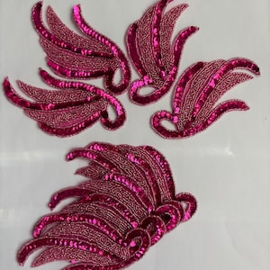 May include: A collection of five fuchsia-colored, sequined and beaded appliques in a feather or wing design. The appliques are arranged on a white surface, showcasing their intricate details and vibrant color.