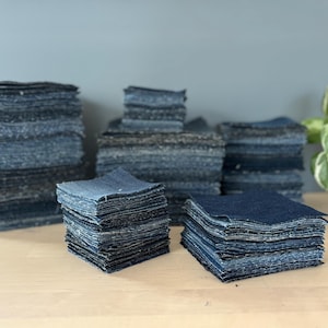 May include: Multiple stacks of square denim fabric patches in various shades of blue, ranging from light to dark indigo. The fabric pieces are neatly piled in several groups of varying heights, showcasing the textured weave of the cotton material.