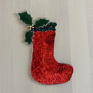 May include: A red sequined Christmas stocking with a silver beaded border. The stocking is decorated with a green holly leaf and berry design. A festive holiday decoration.