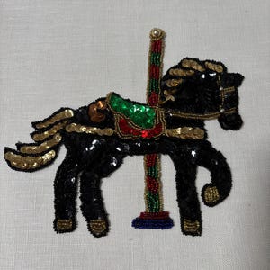 May include: A black sequined carousel horse with gold and green accents. The horse has a red, green, and gold saddle and a multi-colored pole. The horse is embellished with gold sequins and beads.