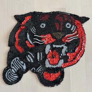 May include: A black beaded tiger head patch with red sequin accents. The tiger's face features red and white details, with gray and gold beadwork. The patch is designed with a fierce expression and detailed beadwork.