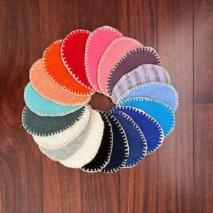 May include: A circular arrangement of multiple felt fabric pieces in various colors including red, orange, pink, purple, blue, green, black, white, and gray. Each piece is shaped like a rounded petal and features a cream-colored blanket stitch.