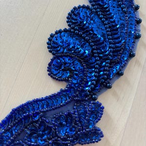 May include: A decorative applique featuring vibrant royal blue sequins and matching beads arranged in a curved, organic pattern on a sheer mesh base. The design consists of layered, fan-like shapes outlined with small, dark blue beads.