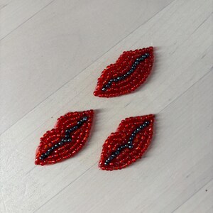May include: Three embroidered patches shaped like lips, crafted with vibrant red seed beads forming the outer shape and dark metallic beads creating a central line to represent the mouth opening. Each patch features a distinct, textured beaded surface.