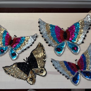 May include: Four butterfly-shaped patches decorated with sequins and beads. Three patches feature a vibrant pattern of silver, gold, turquoise, and magenta sequins with black beaded bodies. One patch is primarily gold and black with sequin detailing.