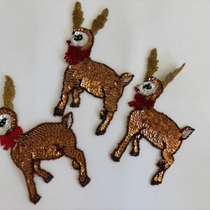 May include: Three decorative reindeer ornaments. Each reindeer is covered in bronze sequins, with gold beaded antlers, a red bow, and white and black facial details. The ornaments are on a white background.