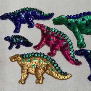 May include: Embroidered dinosaur patches in various colors, including blue, green, pink, and gold. Each patch is covered in sequins and features a spiky ridge along the back. These patches are ideal for adding a decorative touch to clothing or accessories.