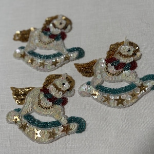 May include: Three decorative rocking horse ornaments. Each horse features a white body adorned with sequins, a gold mane and wings, and a teal base with gold star accents. The horses have red and gold accents around their necks.