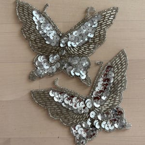 May include: Two silver butterfly appliques. Each butterfly is embellished with silver sequins and beads. The wings have a detailed, textured appearance. The appliques are likely for crafting or embellishing clothing and accessories.