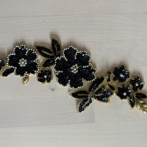May include: A decorative floral applique featuring black sequined flowers with pearl-like centers and gold beaded edges. The applique has several leaves and small crystal accents, creating an elegant embellishment.
