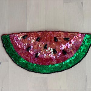 May include: A sequined watermelon slice with a green rind, red and pink flesh, and black seed accents. The decorative piece is made of shiny sequins and is likely used for embellishment or crafting projects.