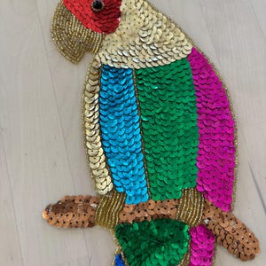 May include: A colorful sequined parrot decoration. The bird features a red head, gold and white body, and blue, green, and pink wings. The parrot is perched on a brown branch and outlined with gold beads.