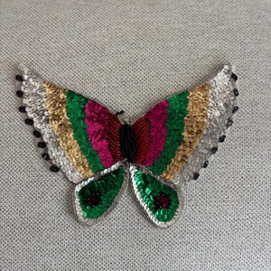 May include: A colorful butterfly applique with sequined wings. The wings feature stripes of silver, gold, green, and pink. The butterfly's body is made of red beads, and the edges of the wings are outlined with black beads.