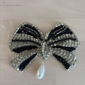 May include: A bow-shaped brooch featuring alternating stripes of silver bugle beads and black sequins. The center is embellished with clear crystals, and a single white teardrop-shaped faux pearl hangs from the bottom of the bow structure.