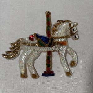 May include: A white carousel horse applique, embellished with sequins and beads. The horse features gold accents, a blue and green saddle, and a red and gold pole. The applique is on a white background.