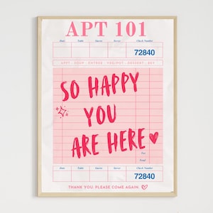 May include: A framed print featuring a pink and white design resembling a restaurant check. The text "APT 101" and "72840" are visible. Bold, red text reads "SO HAPPY YOU ARE HERE" with a heart symbol.