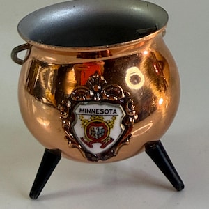 May include: A small copper-coloured cauldron with three black legs and a silver-coloured interior. A decorative shield with the word "MINNESOTA" and a state emblem is affixed to the front. The cauldron has a small handle on one side.