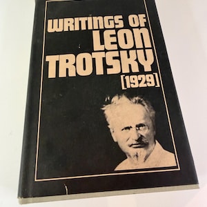 May include: A hardcover book titled "Writings of Leon Trotsky [1929]" with a black cover and tan lettering. The cover features a portrait of Leon Trotsky. The book is on a white surface.