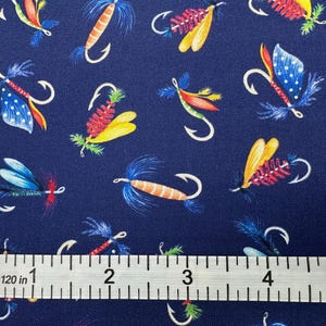May include: Navy blue fabric with a repeating pattern of colorful fishing lures. The lures feature various designs with red, yellow, blue, and orange accents. A ruler shows measurements up to 5 inches.