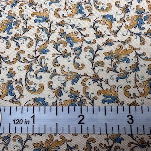 May include: Cream-colored fabric with a repeating pattern of stylized blue and gold floral elements. A ruler shows measurements in inches.