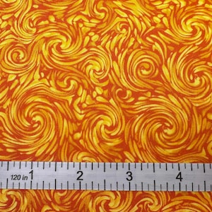 May include: Close-up of a fabric with a swirling pattern in shades of orange and yellow. A metal ruler with inch measurements is placed across the fabric, showing the scale. The fabric design features a repeating, abstract, spiral motif.