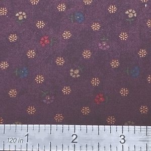 May include: Close-up of a fabric with a dark purple background. The fabric is patterned with small, gold-colored starbursts and tiny flowers in red, blue, and purple. A metal ruler with inch measurements is at the bottom.