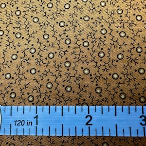 May include: Close-up of a fabric with a repeating pattern of black branches and white-centered circles on a brown background. A blue measuring tape shows the fabric's dimensions, with the 1-inch mark visible.