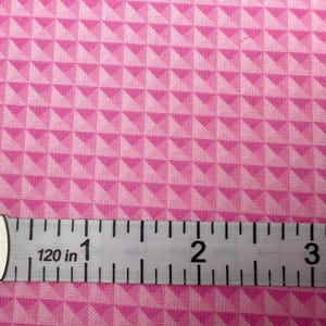 May include: Pink fabric with a geometric pattern of shaded triangles. A white and black measuring tape shows measurements in inches. The fabric has a repeating pattern of light and dark pink triangles.