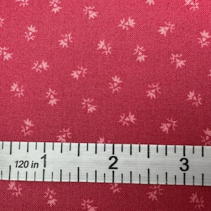 May include: Close-up of a red fabric with a small, repeating floral pattern in a lighter shade of red. A white measuring tape shows 3 inches. The fabric has a textured appearance.