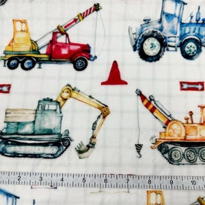 May include: White fabric featuring watercolor illustrations of construction vehicles, including a red crane truck, a blue tractor, and an excavator. Red traffic cones and directional arrows are also scattered across the fabric.