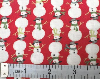 May include: A red fabric featuring a repeating pattern of white snowmen wearing grey hats and green scarves, positioned above a measuring tape that shows a scale of 4 inches, with the snowmen arranged in vertical rows across the material.