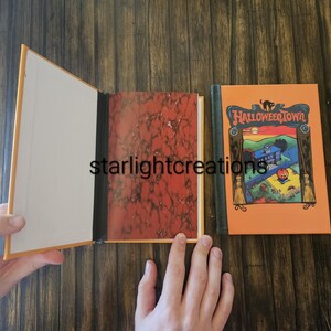 K&ouml;nnte beinhalten: Ein offenes Buch mit cremefarbenem Innenraum und einem marmorierten rot-orangen Muster auf der rechten Seite. Der Einband des Buches auf der rechten Seite ist orange mit den Worten "Halloween Town" und einer farbenfrohen Illustration eines Hauses und eines K&uuml;rbisses.