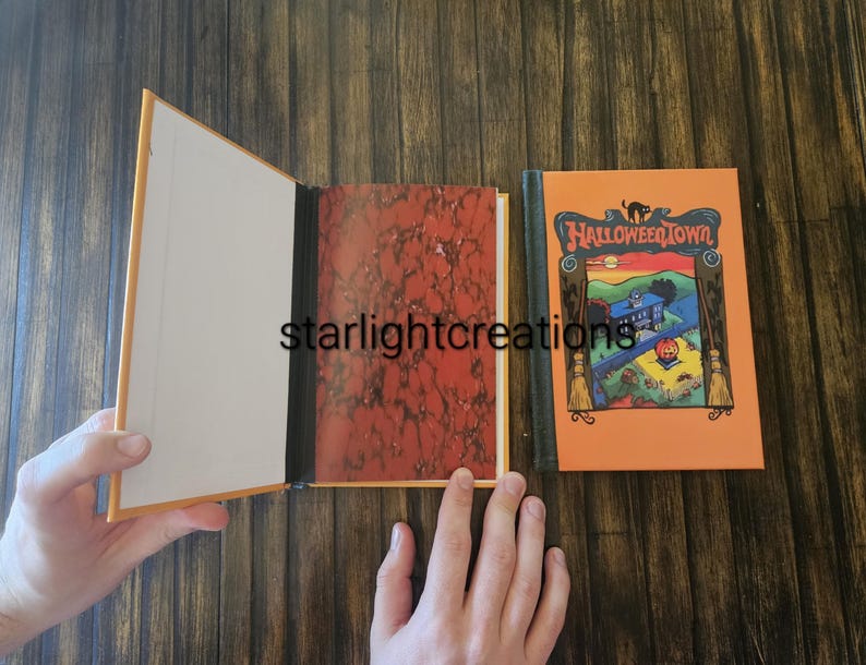 K&ouml;nnte beinhalten: Ein offenes Buch mit cremefarbenem Innenraum und einem marmorierten rot-orangen Muster auf der rechten Seite. Der Einband des Buches auf der rechten Seite ist orange mit den Worten "Halloween Town" und einer farbenfrohen Illustration eines Hauses und eines K&uuml;rbisses.
