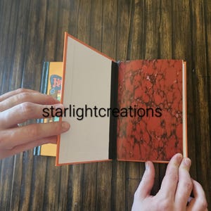 K&ouml;nnte beinhalten: Ein offenes Buch mit orangefarbenen Einb&auml;nden und einem marmorierten rot-braunen Muster auf der rechten Seite. Die linke Seite ist leer. Der Text "starlightcreations" ist sichtbar. Ein weiteres Buch ist im Hintergrund teilweise sichtbar.