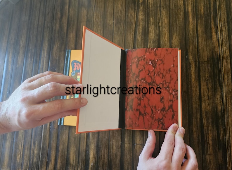 K&ouml;nnte beinhalten: Ein offenes Buch mit orangefarbenen Einb&auml;nden und einem marmorierten rot-braunen Muster auf der rechten Seite. Die linke Seite ist leer. Der Text "starlightcreations" ist sichtbar. Ein weiteres Buch ist im Hintergrund teilweise sichtbar.