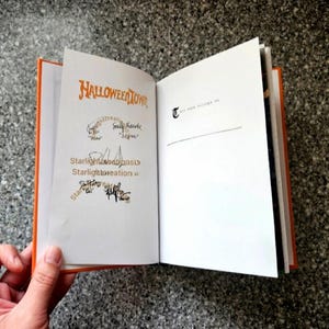 K&ouml;nnte beinhalten: Ein offenes Buch mit orangefarbenen Kanten und wei&szlig;en Seiten. Die linke Seite zeigt den Titel "Halloweentown" in orangefarbener Schrift, mit Unterschriften und dem Text "Starlight Creations". Die rechte Seite hat den Text "T" und "This book belongs to me".
