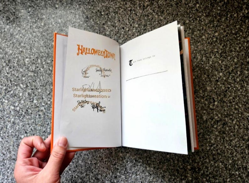 K&ouml;nnte beinhalten: Ein offenes Buch mit orangefarbenen Kanten und wei&szlig;en Seiten. Die linke Seite zeigt den Titel "Halloweentown" in orangefarbener Schrift, mit Unterschriften und dem Text "Starlight Creations". Die rechte Seite hat den Text "T" und "This book belongs to me".